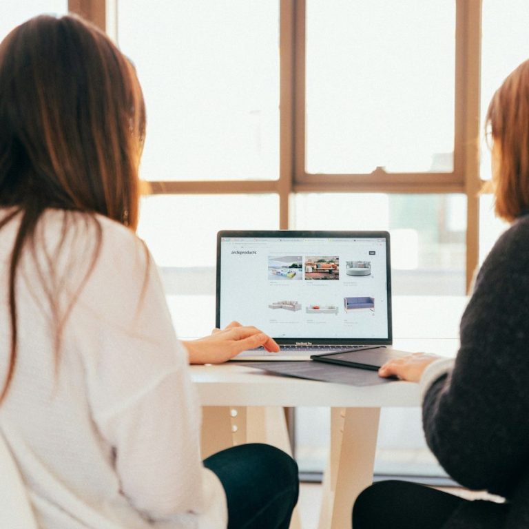 Two people working on a laptop at a table, with large windows in the background.